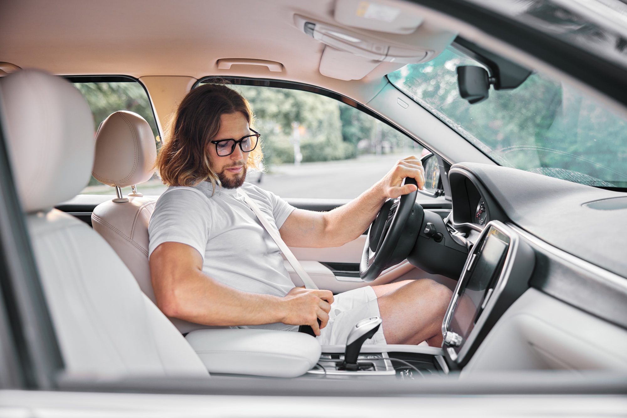 A man in a light shirt sits in the driver s seat of a modern car, adjusting his seatbelt, focused on